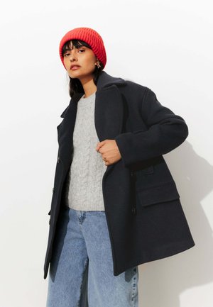 Manteau en mélange de laine bleu marine avec double boutons et poches avant, superposé à un pull en maille torsadée gris clair, associé à un jean ample bleu et un bonnet rouge.