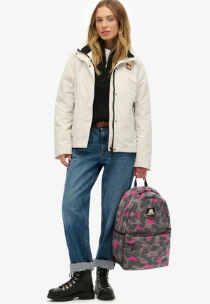 Mochila de camuflaje gris con acentos en rosa, fabricada en un material duradero, con cierre de cremallera y un bolsillo frontal, combinada con una chaqueta blanca y un suéter negro.