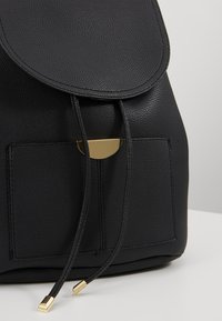 Sac à dos en cuir noir texturé avec poche avant, fermeture à rabat, sangles à cordon et accents métalliques dorés sur les cordons et le rabat.