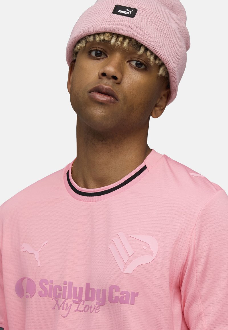Camiseta deportiva de color rosa claro con mangas cortas, con un cuello negro y logotipos en tonos más oscuros. Se lleva con un gorro de punto rosa.