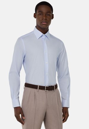 Camisa de botones a rayas azul claro con cuello, mangas largas y un diseño ajustado, combinada con pantalones de tela beige claros con pliegues y un cinturón oscuro.