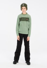 Žalias vilnonis džemperis su juodomis juostelėmis ir logotipu, derinamas su juodomis kelnėmis ir žaliu kepurėliu. Lygi tekstūra, laisvas siluetas.