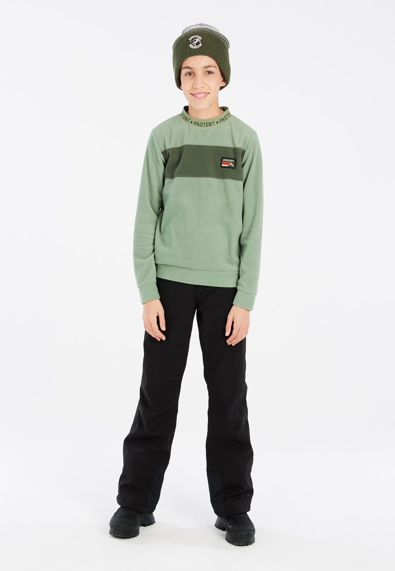 Žalias vilnonis džemperis su juodomis juostelėmis ir logotipu, derinamas su juodomis kelnėmis ir žaliu kepurėliu. Lygi tekstūra, laisvas siluetas.