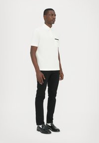 Hvit polo-skjorte med en svart lomme, kombinert med svarte slanke bukser og svarte loafers. Modellen står foran en nøytral bakgrunn.