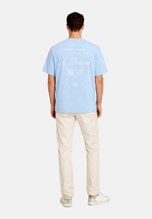 Man wearing light blue T-shirt with white "Le Parisien" and tennis design on back, beige pants, and white sneakers, standing facing away.