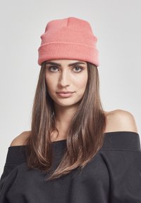 Rosa strikket lue med ribbestruktur, med en midtseam, båret med en svart av-shoulder sweatshirt. Lange, rette brune hår er synlige.