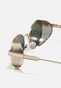 Lunettes de soleil rondes avec une monture en métal doré, des verres verts et des panneaux latéraux perforés. Elles comprennent des plaquettes de nez ajustables et des branches élégantes.