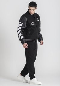 Čierna varsity bunda s koženými rukávmi, bielymi pruhmi a vyšitým logom. Spárovaná s čiernymi nohavicami a bielymi teniskami.
