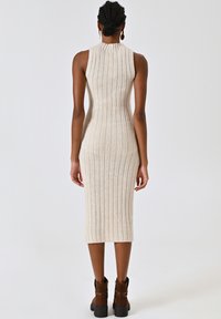 Abito midi in maglia di un beige chiaro, con texture a costine e design senza maniche. L'orlo termina leggermente sotto il ginocchio. Abbinato a stivaletti alla caviglia marroni.