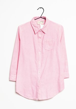 Jasnoróżowa koszula z kołnierzykiem i guzikami wyeksponowana na czarnym wieszaku. Posiada długie rękawy, kieszeń na piersi oraz zaokrąglony dół.