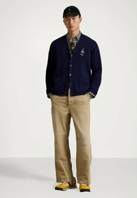 Man med svart keps, marinblå cardigan över rutiga skjortan, beige byxor och gula sneakers, står med händerna i fickorna mot vit bakgrund.