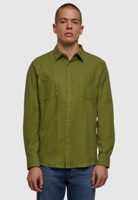 Camisa de botones verde oliva con cuello, dos bolsillos en el pecho, mangas largas y un tejido ligeramente texturizado. Combinada con vaqueros azules.