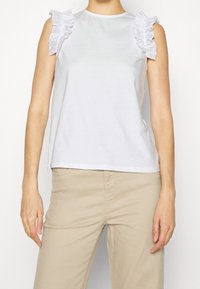 Biała top bez rękawów z falbaniastymi akcentami na ramionach, wykonany z gładkiego materiału. Prosty krój, zestawiony z beżowymi spodniami.