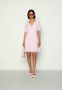 Mulher vestindo um vestido rosa claro de manga curta com decote em V, sandálias brancas, óculos de sol rosas, e segurando uma pequena bolsa bege, de pé contra um fundo simples.