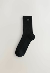 Schwarze gerippte Socken mit einer glatten Textur und einem dekorativen weißen Schleifenemblem nahe der Oberkante. Mid-Calf-Länge.
