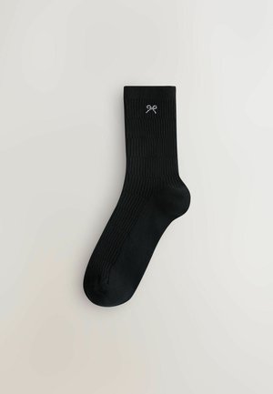 Schwarze gerippte Socken mit einer glatten Textur und einem dekorativen weißen Schleifenemblem nahe der Oberkante. Mid-Calf-Länge.