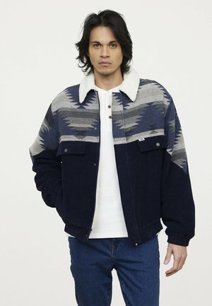 Marineblauwe en grijze corduroyjas met een sherpa-kraag, voorzien van een geometrisch patroon en twee borstzakken. Model draagt een witte henley en jeans.