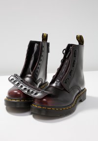 Bottes en cuir noir et rouge foncé avec des coutures jaunes, des fermetures éclair frontales partiellement ouvertes et des panneaux d'œillets amovibles sur une surface blanche.