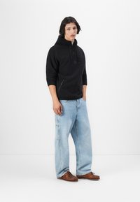 Felpa nera con cappuccio e tasca a marsupio, indossata con jeans larghi azzurri e scarpe da barca in pelle marrone. Design semplice e casual.