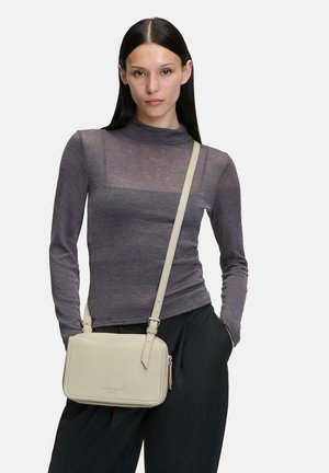 Helle graue Umhängetasche aus glattem Leder, mit einem Reißverschlusstasche und verstellbarem Träger. Das Modell trägt ein enges, dunkelgraues Langarmoberteil.