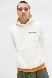 Roomkleurige hoodie met een voorkantzak en zwarte tekst. Gelaagd met een oranje shirt eronder, gedragen met olijfgroene broek.