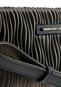 Bolso de tela plisada en gris oscuro con un acabado texturizado. Presenta un acento de hardware negro con una placa de logo plateada. Detalle de correa de cuero visible.