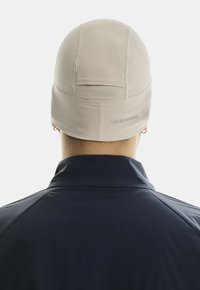 Bonnet en tricot beige clair avec des détails de couture et "ON RUNNING" imprimé en gris à l'arrière. Porté avec une veste bleu foncé, vu de l'arrière.