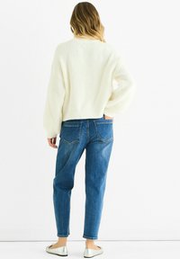 White ribbed sweater with oversized fit, paired with blue denim jeans featuring back pockets and subtle stitching. Model wears silver flats.
