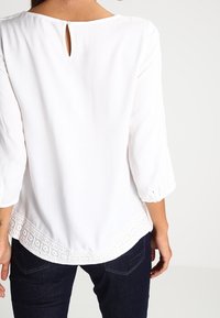 Blusa bianca con apertura a chiave sul retro, maniche a tre quarti e orlo decorativo in pizzo. Tessuto morbido con una consistenza liscia.