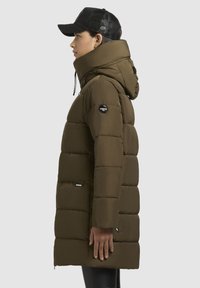 Oliven grøn pufferjakke med høj krave og justerbar hætte. Har en lynlåslomme og logo-patch på den øverste arm. Strukturert stof.
