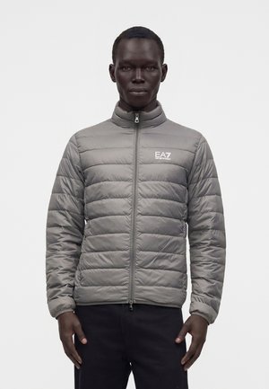 Mężczyzna w jasnoszarej pikowanej kurtce na zamek z logo EA7 Emporio Armani na lewej piersi, stojący na jednolitym jasnym tle.