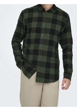 Mężczyzna w zielono-czarnej koszuli w kratę z długim rękawem i beżowych spodniach, z jedną ręką w kieszeni, na jednolitym tle.