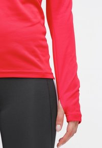 Langærmet atletisk top i lys pink med tekstureret stof, pareret med sorte leggings. Har tommelhulsmuffe for ekstra dækning.