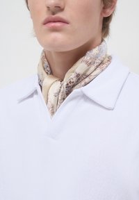 Foulard di seta, beige con intricate fantasie floreali marroni e blu, annodato attorno al collo. Indossato con una polo bianca testurizzata.