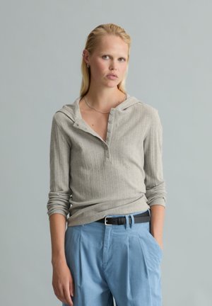 Blondynka w szarej, długorękawowej koszulce henley z guzikami i jasnoniebieskich spodniach z czarnym paskiem, stojąca na jednolitym tle.