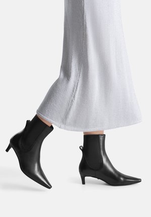 Czarne skórzane botki za kostkę z ostrym noskiem, niskim obcasem typu kotek i elastycznymi panelami po bokach, zestawione z błyszczącą jasnoszarą spódnicą.