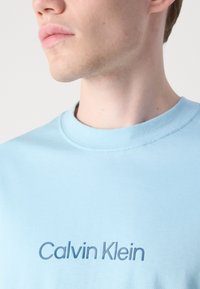 Lyseblå bomull T-skjorte med rund hals og brodert "Calvin Klein"-logo i mørkere blå. Glatt tekstur med korte ermer.