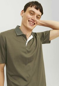 Olivovo zelená polokošeľa s textúrovaným šedým golierom, bielym akcentom a detailom loga. Krátke rukávy, bavlnený materiál, hladká textúra.