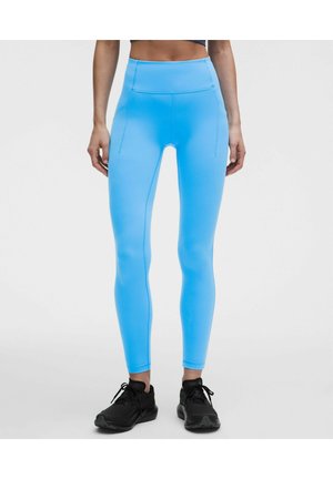 Person iført lyseblå højtaljede leggings og sorte sportssko står mod en ensfarvet hvid baggrund.