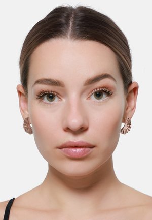 Boucles d'oreilles clou en or rose rainuré avec un design en forme de haricot, présentant une finition polie et des accents texturés, mettant en valeur le visage de la personne qui les porte.