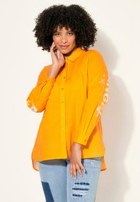 Camicetta arancione brillante con colletto, maniche lunghe e testo grafico bianco con design di stelle sulla manica. Indossata con jeans chiari strappati.