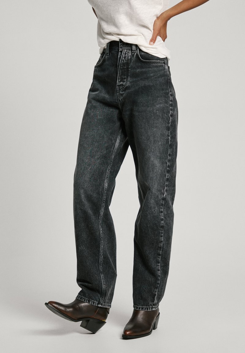 Aukštos juosmens juodi džinsai su tiesiomis kojomis ir subtiliu nudeginimu. Suderinti su rudos spalvos aukštesniais batais, kurie demonstruoja klasikinį dizainą.