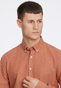 Camicia color ruggine, con bottoni e colletto, realizzata in tessuto testurizzato. I bottoni bianchi contrastano con la tonalità del tessuto.
