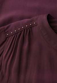 Burgundská blůza z měkké látky, se skládací částí a lemem zdobeným malými kovovými hroty podél ramenního švu.