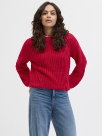 Röd stickad tröja med strukturerad stickning, rund hals och lös passform, tillsammans med ljusblå raka jeans mot en neutral vit bakgrund.