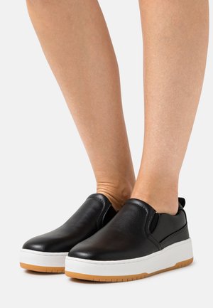 Sapatos pretos tipo slip-on feitos de couro liso, com uma sola branca e um detalhe em borracha de guisado. Bico arredondado e costuras mínimas.