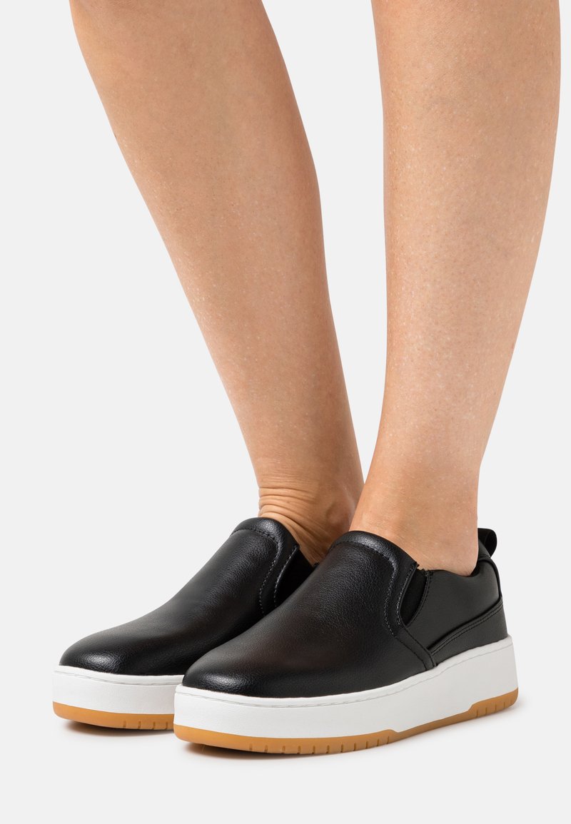Sapatos pretos tipo slip-on feitos de couro liso, com uma sola branca e um detalhe em borracha de guisado. Bico arredondado e costuras mínimas.