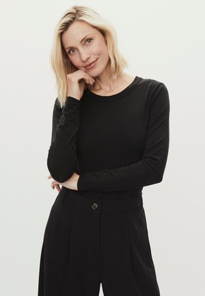 Mujer rubia con cabello corto, vistiendo una blusa negra de manga larga y pantalones negros de talle alto, posando con la mano apoyada en el mentón.