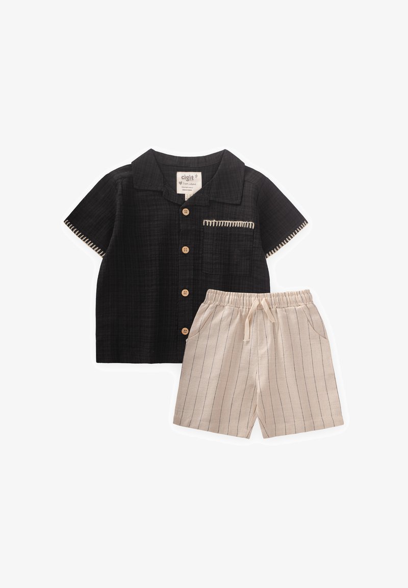 Juodas trumpomis rankovėmis marškinėliai su mediniais mygtukais ir kišene, derinami su smėliniais šortais su vertikaliomis juostelėmis ir elastinga juosmens juosta.