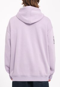 Haut à capuche violet au fit décontracté, avec un cordon de serrage et des poignets côtelés, fabriqué à partir d'un matériau doux. Détail de design notable sur la manche gauche.